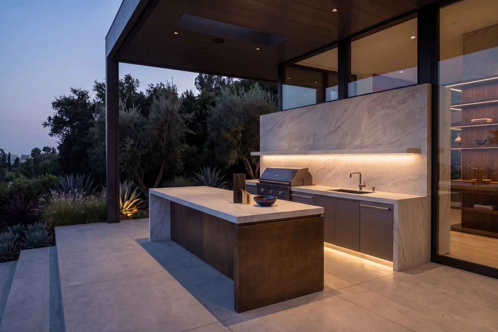 A luxury outdoor kitchen at dusk, sintered stone used as countertop and wall cladding behind a built-in grill, warm lighting illuminating the marble-veined surface against mature Mediterranean landscaping and dusk sky.